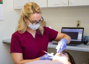 Dental hygienist in scrubs and face mask cleaning a patient's teeth in a dental chair