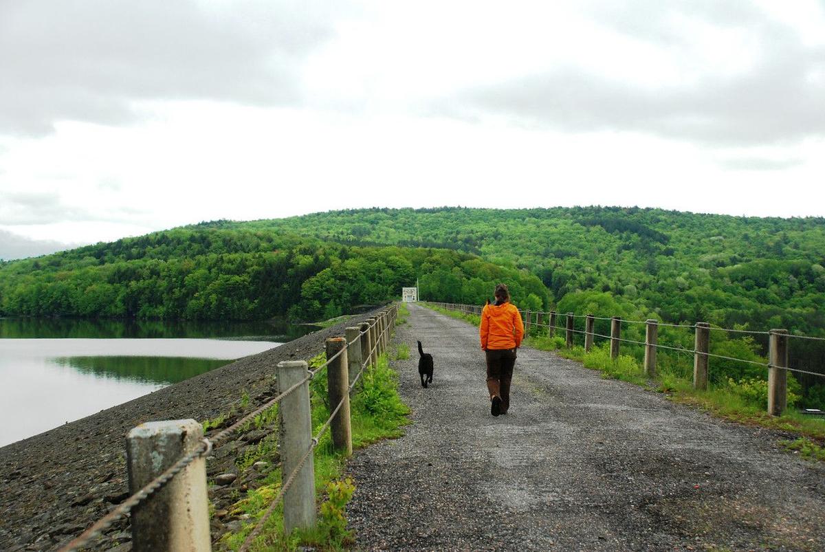 A woman and her dog walk on the gravel path atop a dam by a lake.