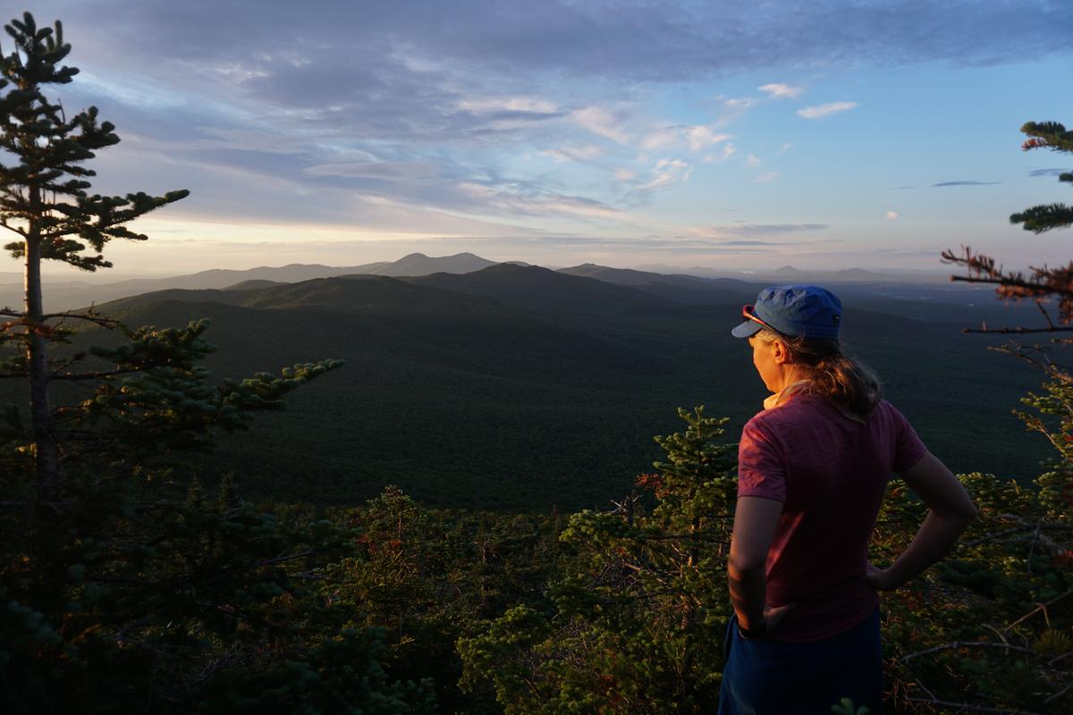 A hiker with a blue hat and gray ponytail stands with their back to the camera, looking out over a colorful sky